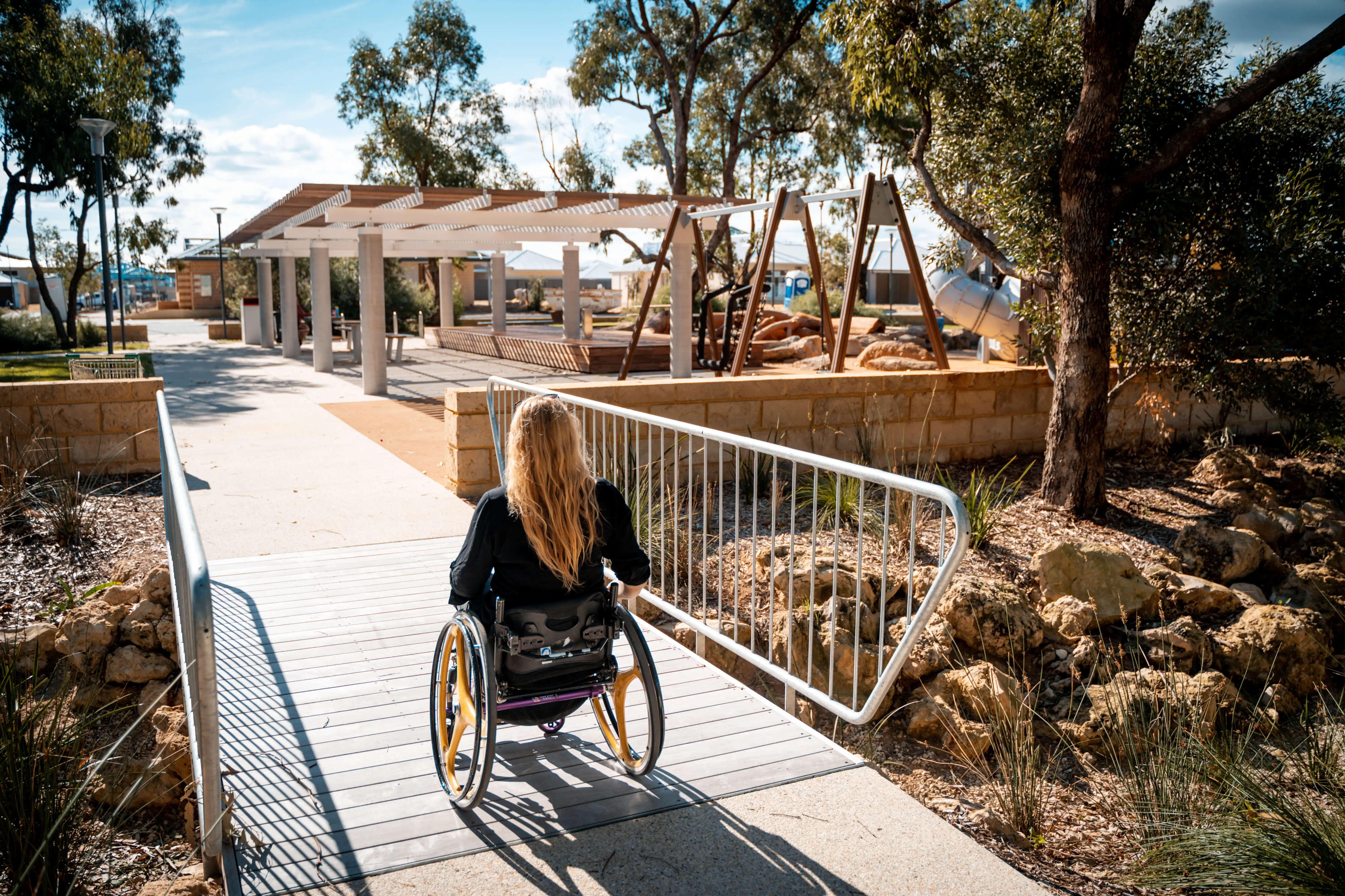 A person using a wheelchair moves along an accessible pathway toward a shaded outdoor area.